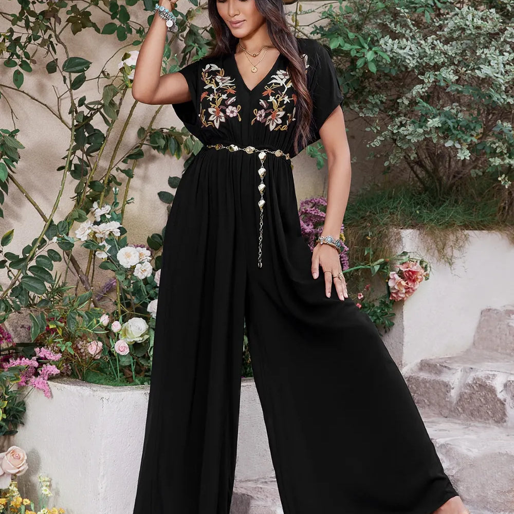 Woman wearing a black boho wide-leg jumpsuit with floral embroidery, gold waist chain, and straw hat, standing on stone steps surrounded by flowers.