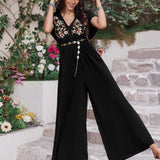 Woman wearing a black boho wide-leg jumpsuit with floral embroidery, gold waist chain, and straw hat, standing on stone steps surrounded by flowers.