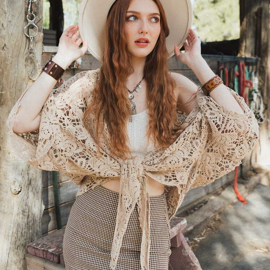 Woman wearing a beige crochet tie-front kimono over a white top and checkered mini skirt, accessorized with a hat and bracelets.