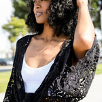 Close-up of woman in black crochet kimono wrap over a white crop top, outdoors in sunlight.