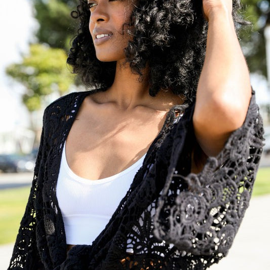 Close-up of woman in black crochet kimono wrap over a white crop top, outdoors in sunlight.