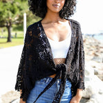 Close-up of woman in black crochet kimono wrap over a white crop top, outdoors in sunlight.