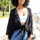 Close-up of woman in black crochet kimono wrap over a white crop top, outdoors in sunlight.