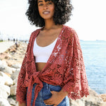 Woman wearing a rust red crochet kimono wrap tied at the front, paired with jeans and a white crop top by the water