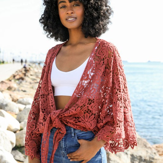 Woman wearing a rust red crochet kimono wrap tied at the front, paired with jeans and a white crop top by the water