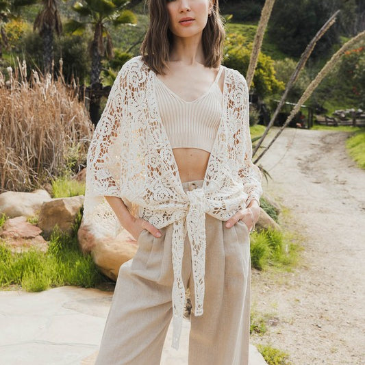 Woman wearing a white floral crochet kimono wrap with wide sleeves, viewed from the back in a tropical outdoor setting.