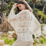 Woman wearing a white floral crochet kimono wrap with wide sleeves, viewed from the back in a tropical outdoor setting.