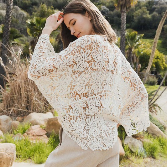 Woman wearing a white floral crochet kimono wrap with wide sleeves, viewed from the back in a tropical outdoor setting.