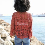 Woman wearing a rust red crochet kimono wrap tied at the front, paired with jeans and a white crop top by the water