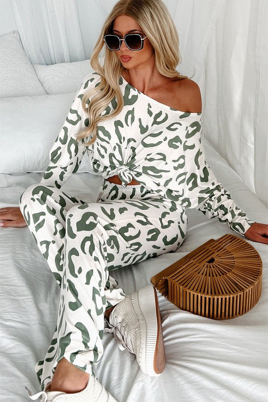 Woman lounging in bed wearing a sage leopard print lounge set with an off-shoulder knotted top and white sneakers.