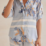 Model wearing a light blue floral lounge set with short-sleeve button-down top and wide-leg pants, featuring peach, navy, and white accents.