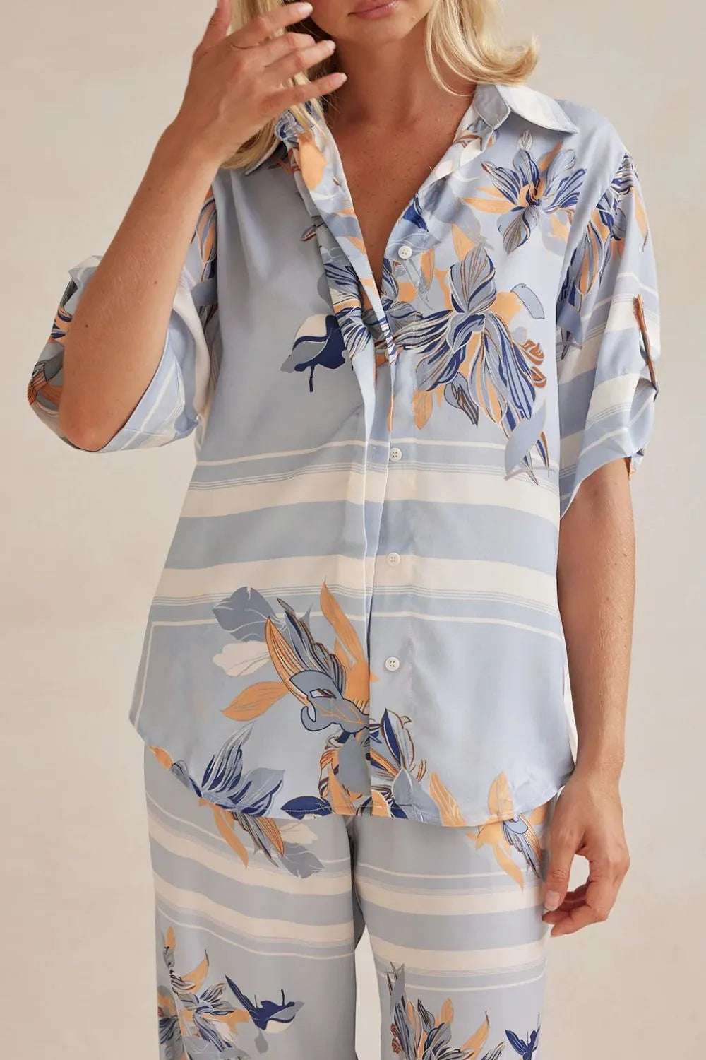 Model wearing a light blue floral lounge set with short-sleeve button-down top and wide-leg pants, featuring peach, navy, and white accents.