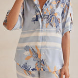 Model wearing a light blue floral lounge set with short-sleeve button-down top and wide-leg pants, featuring peach, navy, and white accents.
