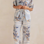 Model wearing a light blue floral lounge set with short-sleeve button-down top and wide-leg pants, featuring peach, navy, and white accents.