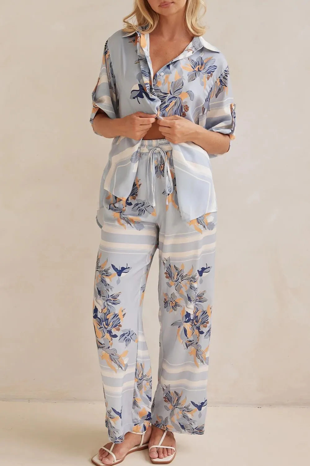 Model wearing a light blue floral lounge set with short-sleeve button-down top and wide-leg pants, featuring peach, navy, and white accents.