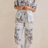 Model wearing a light blue floral lounge set with short-sleeve button-down top and wide-leg pants, featuring peach, navy, and white accents.