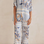 Model wearing a light blue floral lounge set with short-sleeve button-down top and wide-leg pants, featuring peach, navy, and white accents.