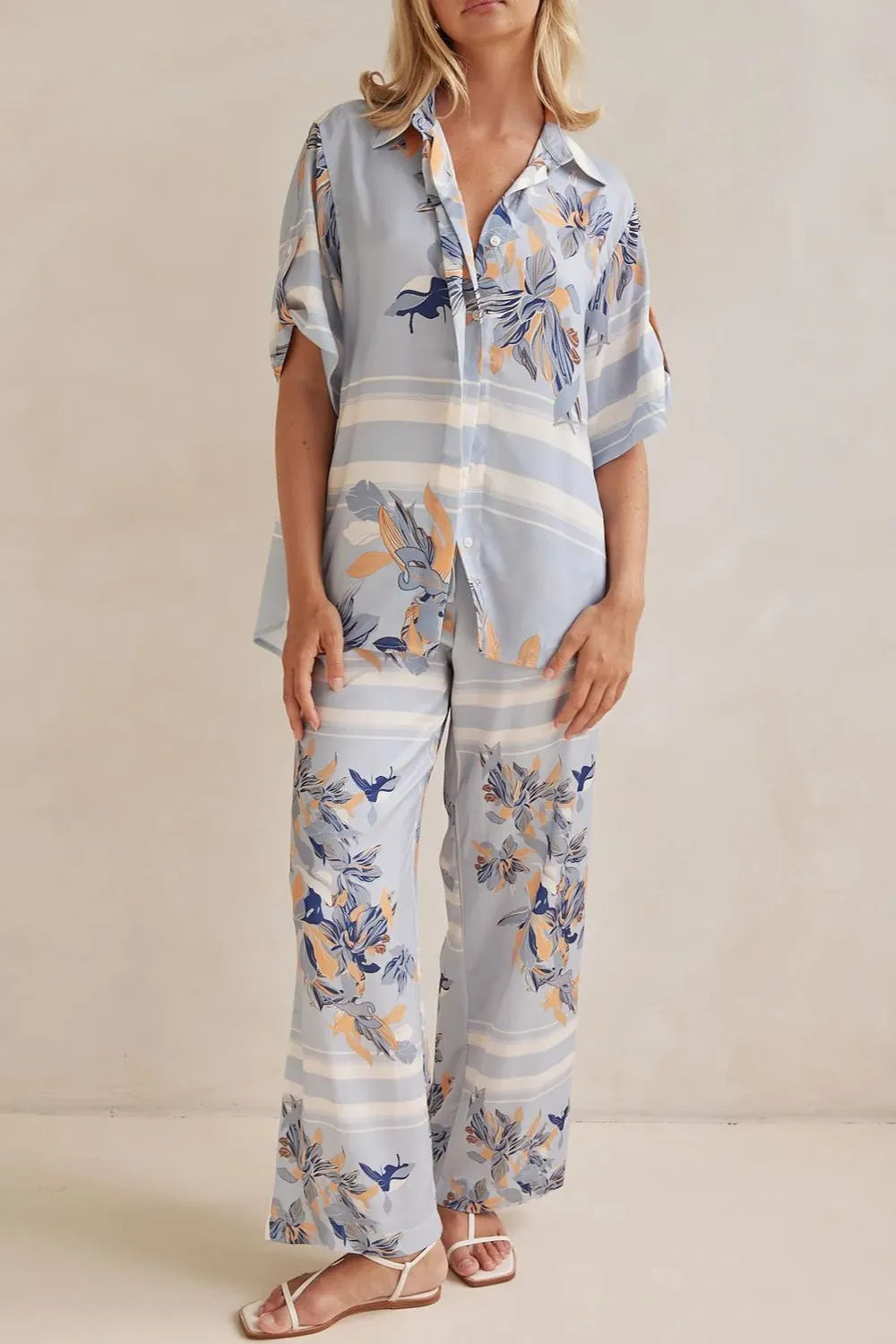 Model wearing a light blue floral lounge set with short-sleeve button-down top and wide-leg pants, featuring peach, navy, and white accents.