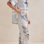 Model wearing a light blue floral lounge set with short-sleeve button-down top and wide-leg pants, featuring peach, navy, and white accents.