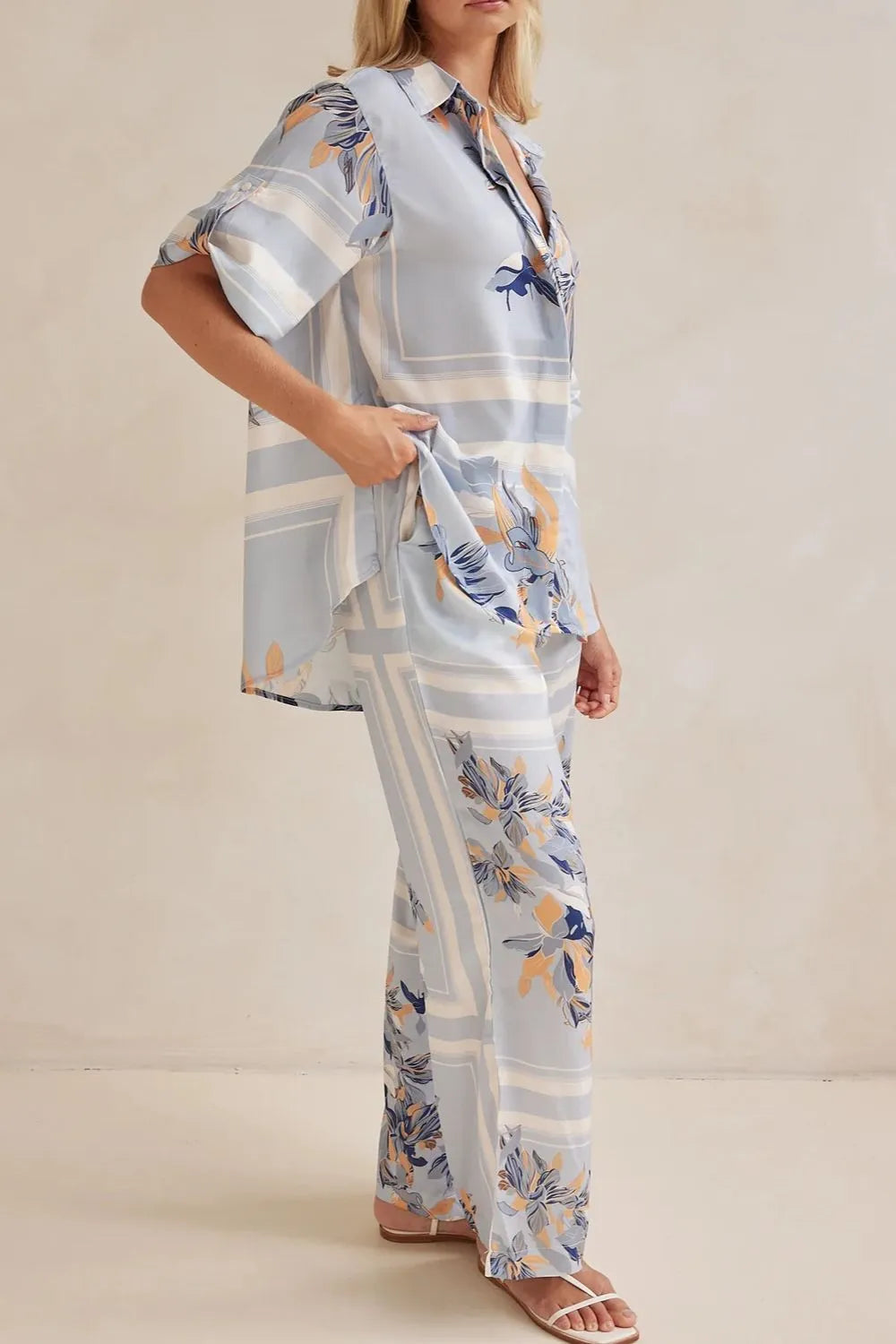 Model wearing a light blue floral lounge set with short-sleeve button-down top and wide-leg pants, featuring peach, navy, and white accents.