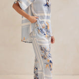 Model wearing a light blue floral lounge set with short-sleeve button-down top and wide-leg pants, featuring peach, navy, and white accents.