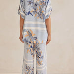 Model wearing a light blue floral lounge set with short-sleeve button-down top and wide-leg pants, featuring peach, navy, and white accents.
