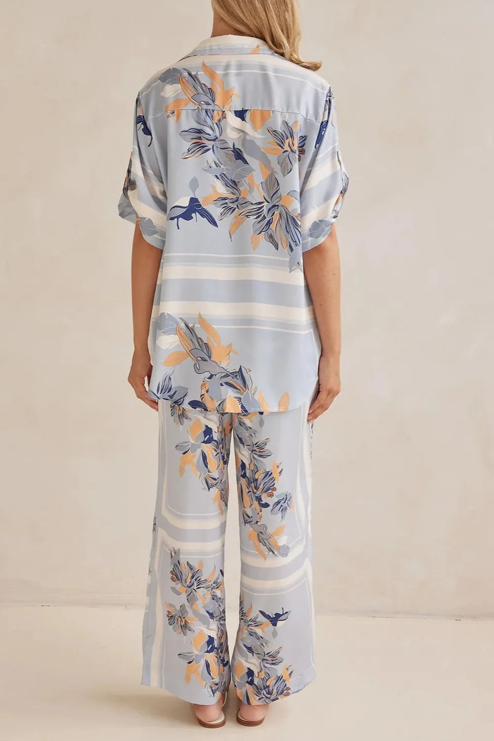 Model wearing a light blue floral lounge set with short-sleeve button-down top and wide-leg pants, featuring peach, navy, and white accents.