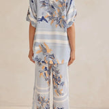 Model wearing a light blue floral lounge set with short-sleeve button-down top and wide-leg pants, featuring peach, navy, and white accents.
