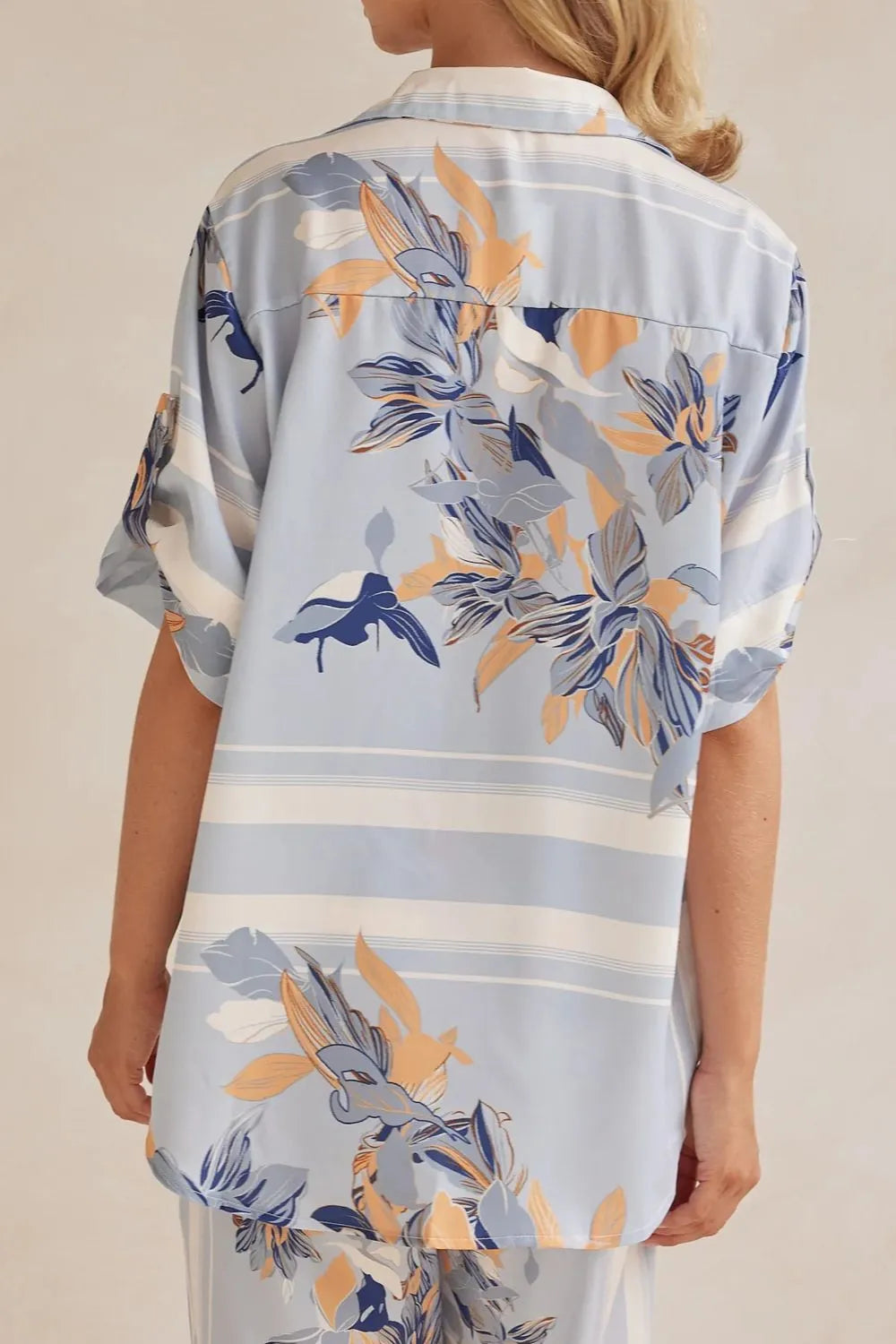 Model wearing a light blue floral lounge set with short-sleeve button-down top and wide-leg pants, featuring peach, navy, and white accents.