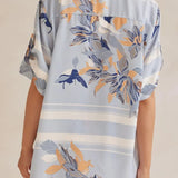 Model wearing a light blue floral lounge set with short-sleeve button-down top and wide-leg pants, featuring peach, navy, and white accents.