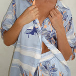 Model wearing a light blue floral lounge set with short-sleeve button-down top and wide-leg pants, featuring peach, navy, and white accents.