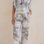 Model wearing a light blue floral lounge set with short-sleeve button-down top and wide-leg pants, featuring peach, navy, and white accents.