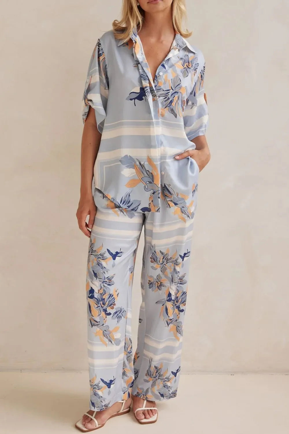 Model wearing a light blue floral lounge set with short-sleeve button-down top and wide-leg pants, featuring peach, navy, and white accents.