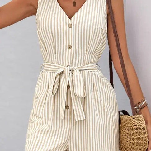 Woman wearing a beige and white vertical striped sleeveless romper with button front and waist tie, styled with hoop earrings and woven bag.