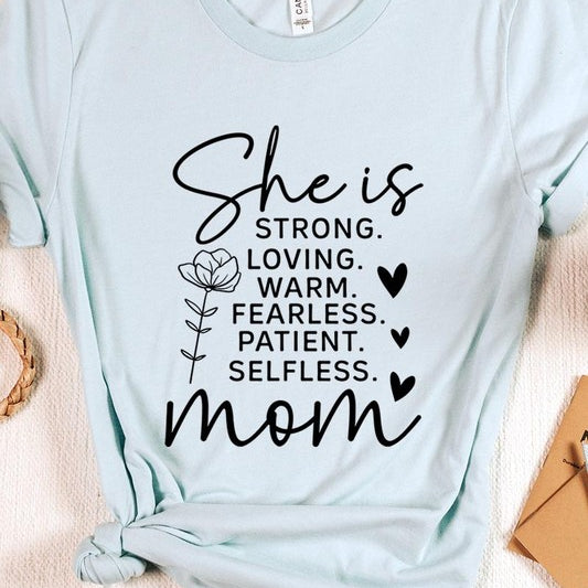 Graphic T-shirt reading ‘She is strong, loving, warm, fearless, patient, selfless—mom’ styled flat-lay with tan fedora, denim shorts, and greenery.