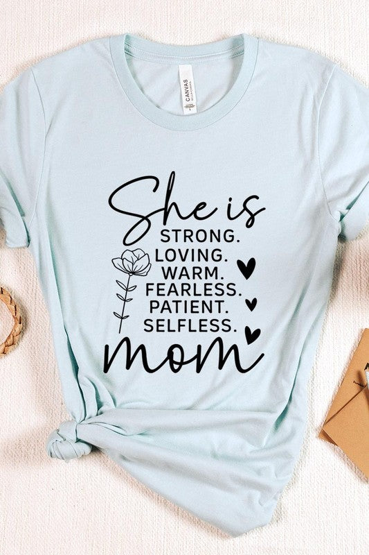 Graphic T-shirt reading ‘She is strong, loving, warm, fearless, patient, selfless—mom’ styled flat-lay with tan fedora, denim shorts, and greenery.