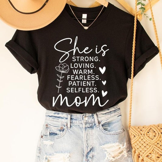 Black graphic T-shirt reading ‘She is strong, loving, warm, fearless, patient, selfless—mom’ styled flat-lay with tan fedora, denim shorts, and greenery.