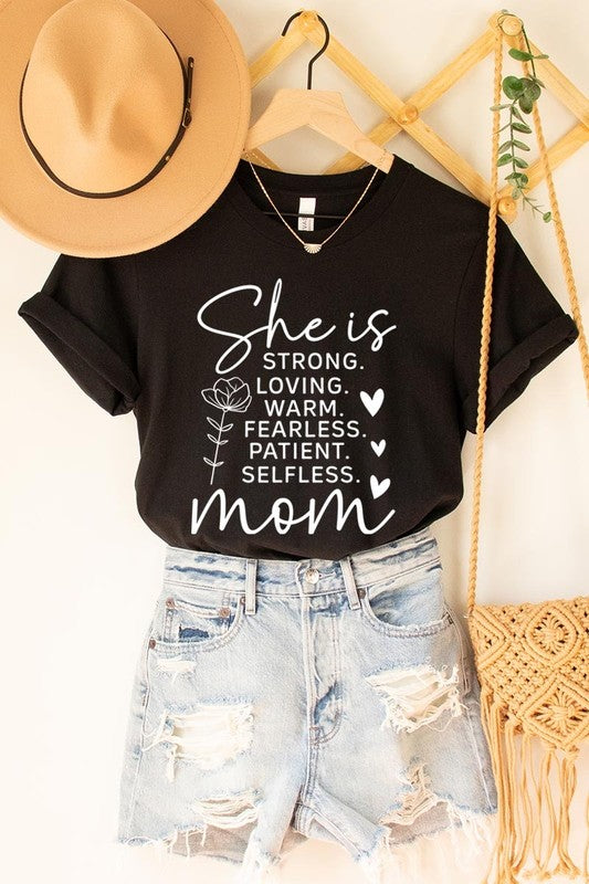 Black graphic T-shirt reading ‘She is strong, loving, warm, fearless, patient, selfless—mom’ styled flat-lay with tan fedora, denim shorts, and greenery.
