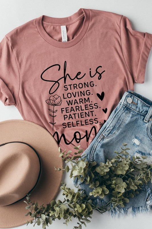 Dusty-rose graphic T-shirt reading ‘She is strong, loving, warm, fearless, patient, selfless—mom’ styled flat-lay with tan fedora, denim shorts, and greenery.