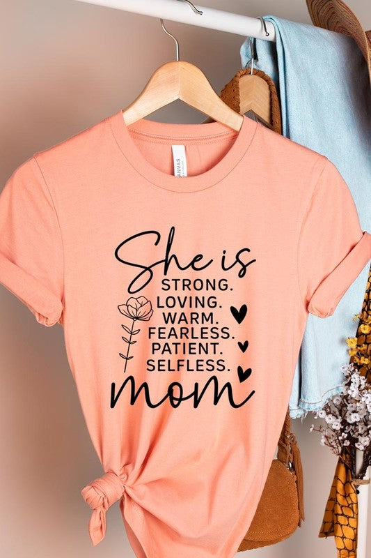 Creamsicle Orange graphic T-shirt reading ‘She is strong, loving, warm, fearless, patient, selfless—mom’ styled flat-lay with tan fedora, denim shorts, and greenery.