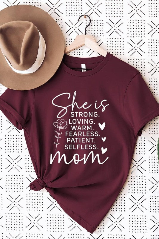 Crimson Red graphic T-shirt reading ‘She is strong, loving, warm, fearless, patient, selfless—mom’ styled flat-lay with tan fedora, denim shorts, and greenery.