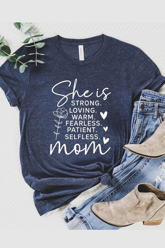 Graphic T-shirt reading ‘She is strong, loving, warm, fearless, patient, selfless—mom’ styled flat-lay with tan fedora, denim shorts, and greenery.