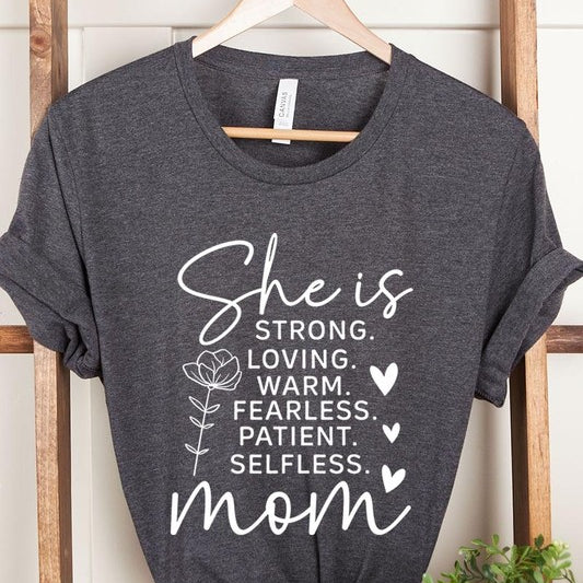 Dark Grey graphic T-shirt reading ‘She is strong, loving, warm, fearless, patient, selfless—mom’ styled flat-lay with tan fedora, denim shorts, and greenery.