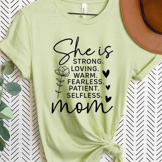 Light Green graphic T-shirt reading ‘She is strong, loving, warm, fearless, patient, selfless—mom’ styled flat-lay with tan fedora, denim shorts, and greenery.