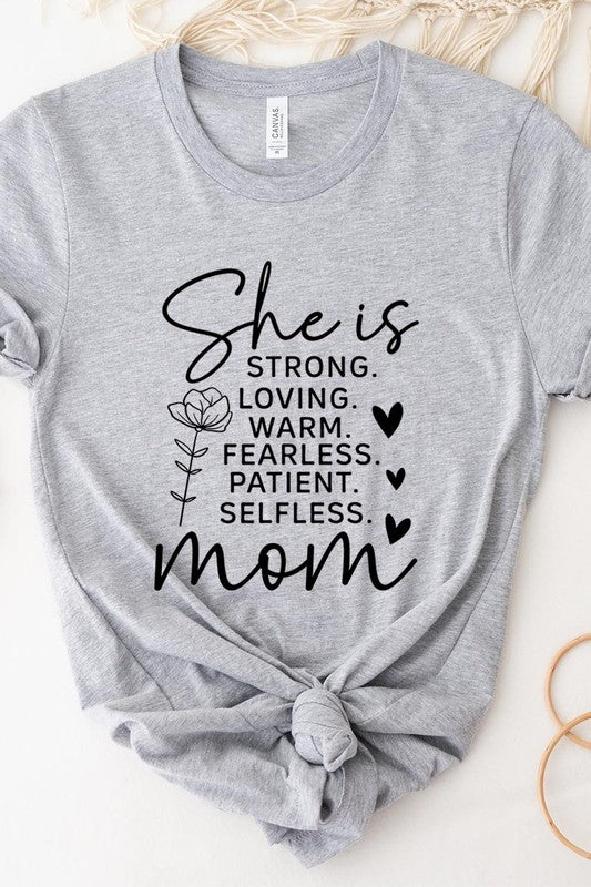 Light Grey graphic T-shirt reading ‘She is strong, loving, warm, fearless, patient, selfless—mom’ styled flat-lay with tan fedora, denim shorts, and greenery.