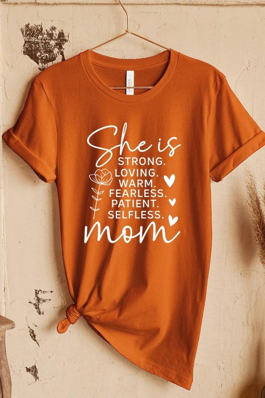 Rusted Orange graphic T-shirt reading ‘She is strong, loving, warm, fearless, patient, selfless—mom’ styled flat-lay with tan fedora, denim shorts, and greenery.