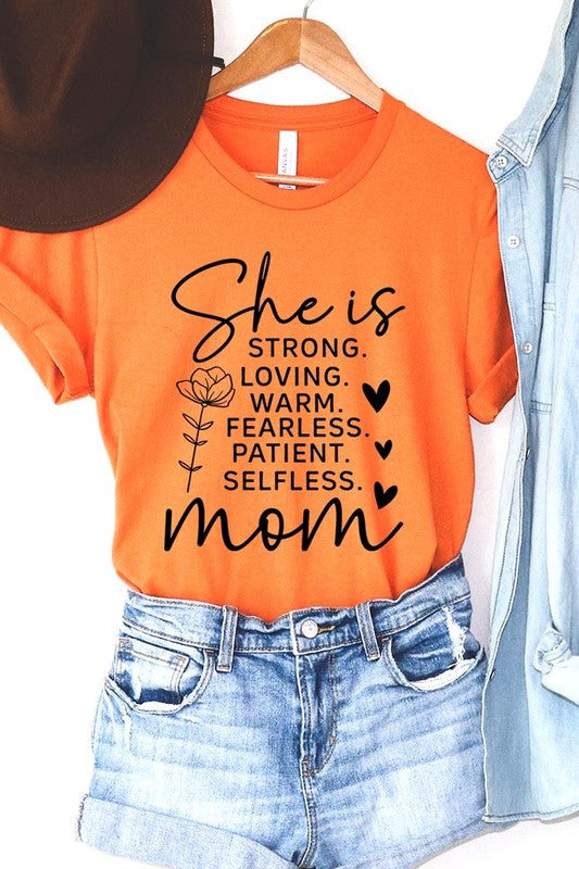 Orange graphic T-shirt reading ‘She is strong, loving, warm, fearless, patient, selfless—mom’ styled flat-lay with tan fedora, denim shorts, and greenery.