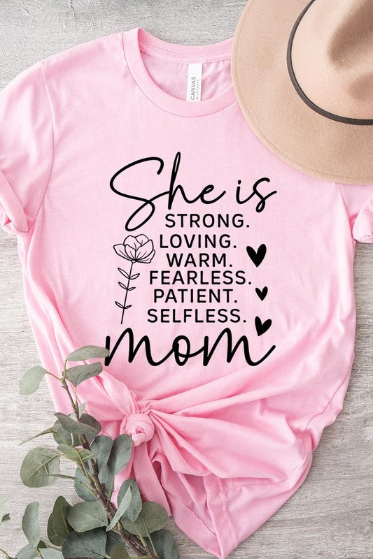 Pink graphic T-shirt reading ‘She is strong, loving, warm, fearless, patient, selfless—mom’ styled flat-lay with tan fedora, denim shorts, and greenery.
