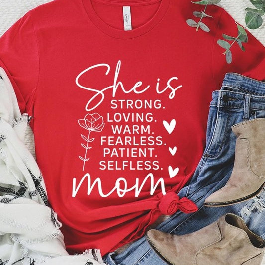Red graphic T-shirt reading ‘She is strong, loving, warm, fearless, patient, selfless—mom’ styled flat-lay with tan fedora, denim shorts, and greenery.