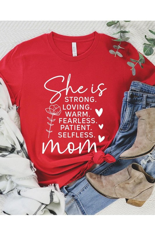 Red graphic T-shirt reading ‘She is strong, loving, warm, fearless, patient, selfless—mom’ styled flat-lay with tan fedora, denim shorts, and greenery.